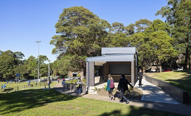 Exterior view of a compact contemporary pavilion or amenity building beside a landscaped public recreation area. The structure has a flat roof, dark brick walls, a deep covered opening, and a concrete pedestrian ramp, with mature trees and sports fields surrounding it.