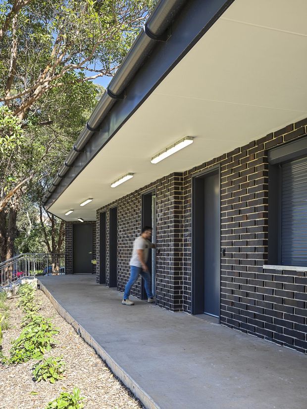Exterior view of a contemporary single-storey building with a long covered concrete walkway. The facade is constructed from dark charcoal brick with contrasting light mortar, dark metal-framed doors and roller shutters, and a broad white soffit fitted with linear fluorescent lights. Dark metal gutters run along the roof edge. A landscaped garden borders the walkway, with trees and railing visible to the side.