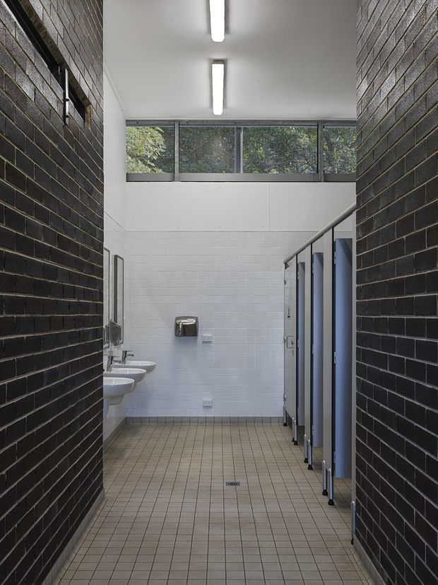 A narrow contemporary public restroom viewed from the entrance, with dark exposed brick walls, white tiled walls, a grid of small beige floor tiles, wall-mounted basins, stainless fixtures, and blue-gray toilet cubicle partitions. High clerestory glazing brings in natural light, complemented by linear fluorescent ceiling lights.