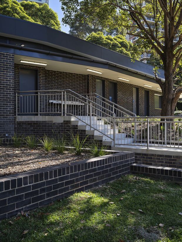 Exterior view of a low-rise institutional or commercial building with dark charcoal brick walls, a broad flat roof canopy, repeated dark doors and narrow windows, and accessible stairs and ramps with silver metal handrails. Raised dark-brick landscaping walls, planted beds, mature trees, and lawn occupy the foreground. A taller apartment or multi-storey building is partly visible behind the trees.