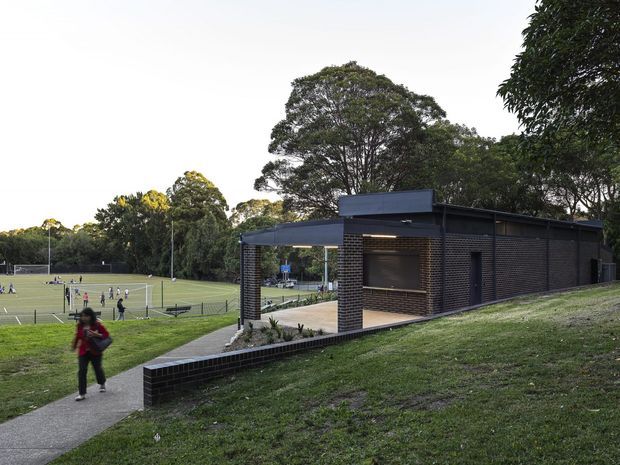A low, modern dark-brick pavilion or amenities building set into a grassy sports-park landscape. The building has a flat roof, covered open-sided entry, exposed dark brick walls, recessed lighting, and a paved pedestrian path leading past it. Sports courts and lush mature trees are visible in the background.