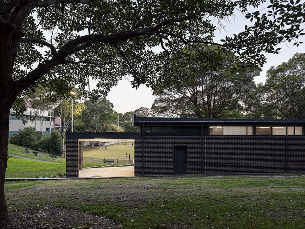 Exterior view of a low, modern dark-brick building set within a landscaped, tree-lined site. The building has a flat roof, long horizontal clerestory-style glazing, dark masonry walls, and a large open passage or covered entry framing the lawn beyond. Mature trees, grass, and a gently sloping campus-like setting surround the structure.