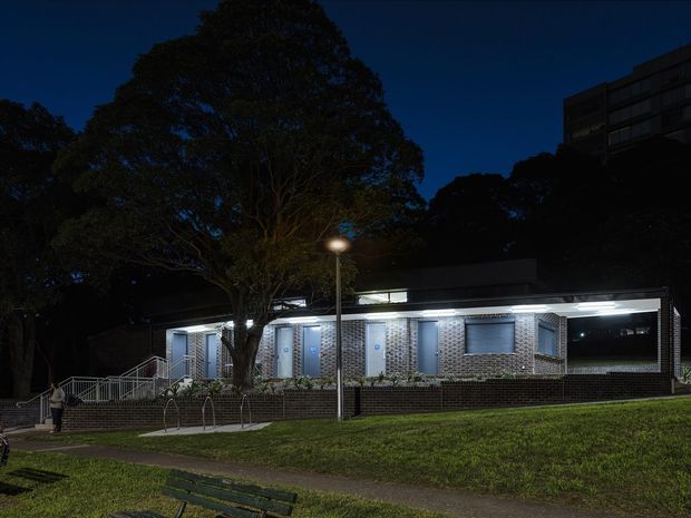 Night-time exterior photograph of a low-rise institutional or commercial building set within a landscaped urban site. The building has a long, rectilinear form with dark brick masonry walls, patterned light brick or blockwork around the windows, large horizontal windows, a flat roof, bright linear exterior lighting, and accessible entry ramps and stairs. Mature trees and a sloping grass verge frame the foreground, while a taller multi-storey building is visible in the background.