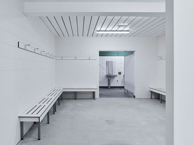 A bright, minimalist commercial changing room or locker-room interior with white painted masonry walls, continuous wall-mounted hooks, simple slatted benches, a smooth grey concrete floor, and a tiled washroom visible through the central opening. The ceiling features white linear slatted panels and fluorescent lighting. The space is functional, clean, and highly utilitarian.