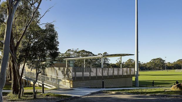 Exterior view of a contemporary low-rise sports or community facility set beside expansive grass playing fields. The building has a pale concrete-block base, glazed or screened upper walls, a broad shallow metal canopy, accessible ramps and railings, and tall light poles. Mature eucalyptus trees and open parkland surround the facility.