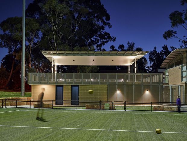 Night-time exterior view of a modern sports facility beside an illuminated artificial turf court. The building has a raised balcony or spectator terrace with metal balustrades, a broad shallow pitched or folded roof canopy, exposed slender columns, and a lower level clad in green masonry block with dark doors and wall-mounted lighting. Trees form a dark backdrop, while blurred players and balls appear on the court.