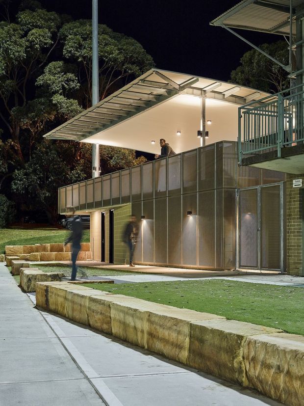 Night-time exterior view of a contemporary multi-level building with a broad translucent roof canopy, exposed steel supports, illuminated covered walkway or terrace, metal screening and railings, and dark brick or masonry walls. The building is set among mature trees and landscaped grounds with sandstone retaining walls and concrete paving. Several blurred pedestrians are visible, suggesting a public or institutional setting.