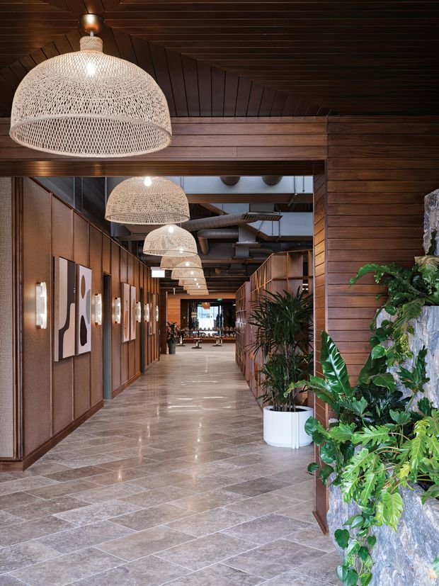 A warmly lit commercial interior hallway with richly stained timber wall and ceiling panelling, polished stone-look tiled flooring, repeated woven pendant lights, framed artwork, built-in cabinetry, and abundant tropical planting. The long corridor leads toward a brighter communal area, creating a refined hospitality or resort-like atmosphere.