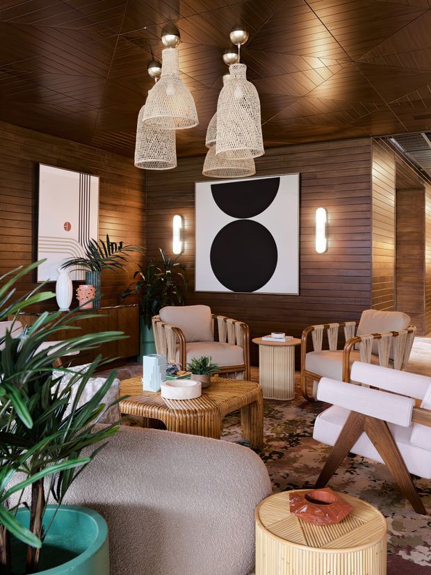 Warm, richly layered lounge interior with dark timber wall and ceiling panelling, woven pendant lights, natural rattan furniture, upholstered armchairs and sofa, abundant indoor plants, graphic artwork, and a patterned rug. The space has a refined tropical and mid-century-inspired character with intimate ambient lighting.