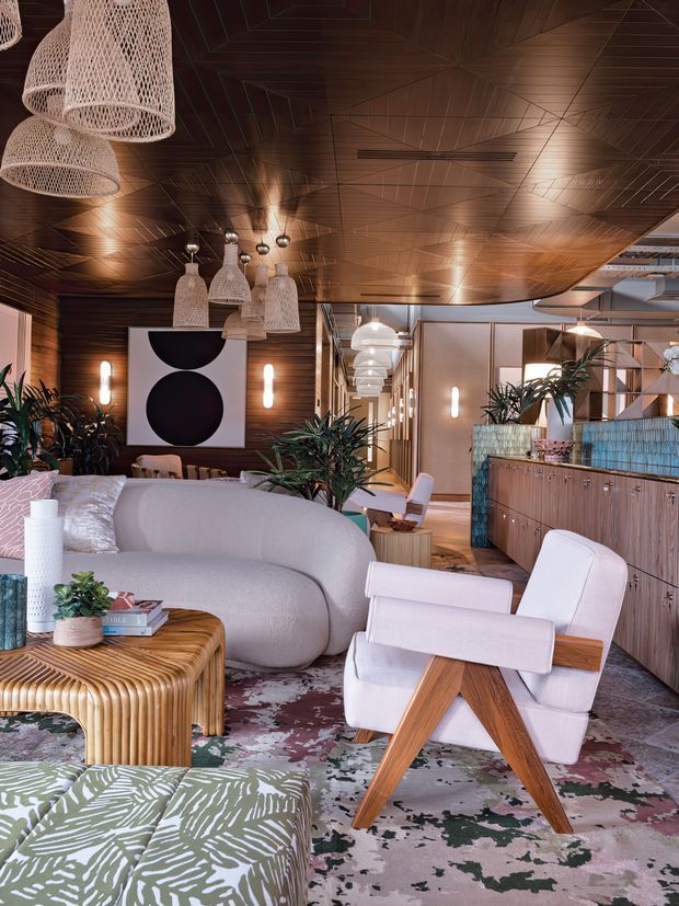 A richly layered contemporary lounge and bar interior with a warm timber-lined ceiling and walls, decorative woven pendant lights, sculptural curved sofa, white and timber lounge chair, ribbed timber coffee table, patterned rug, indoor plants, and a tiled bar counter with timber cabinetry. The space extends into a softly lit corridor with repeated pendant and wall lighting.