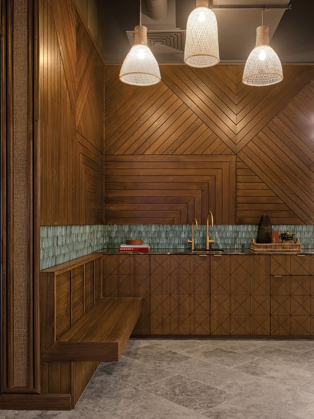 A richly detailed compact kitchen or bar area with extensive warm timber wall panelling arranged in chevron and geometric patterns. A teal-green textured tile splashback runs behind the dark benchtop, with brass-toned tapware and integrated timber cabinetry featuring diamond-pattern fronts. A built-in timber bench occupies the left side, while three woven pendant lights hang from an exposed dark ceiling with visible ductwork. The space has a warm, moody, handcrafted and refined character.