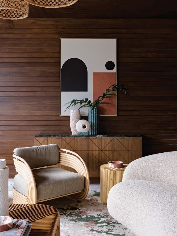 Warm, richly layered living room with dark horizontal timber wall panelling and ceiling, a woven pendant light, sculptural abstract artwork, a timber sideboard with geometric detailing, curved cane-framed upholstered armchairs, a rounded boucle sofa, and a patterned area rug. The room combines natural materials, earthy tones, and organic rounded forms for a refined retro-inspired atmosphere.