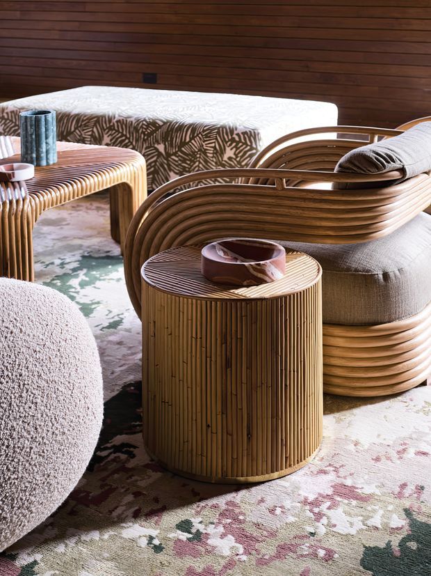 A warmly lit, highly styled living room vignette featuring sculptural curved timber lounge chairs, a cylindrical ribbed timber side table, a rounded upholstered ottoman, and a patterned floral rug. Rich horizontal timber wall panelling forms the backdrop, while a pale patterned upholstered bench and decorative objects add texture and colour. The overall look is organic, tactile, and retro-inspired.
