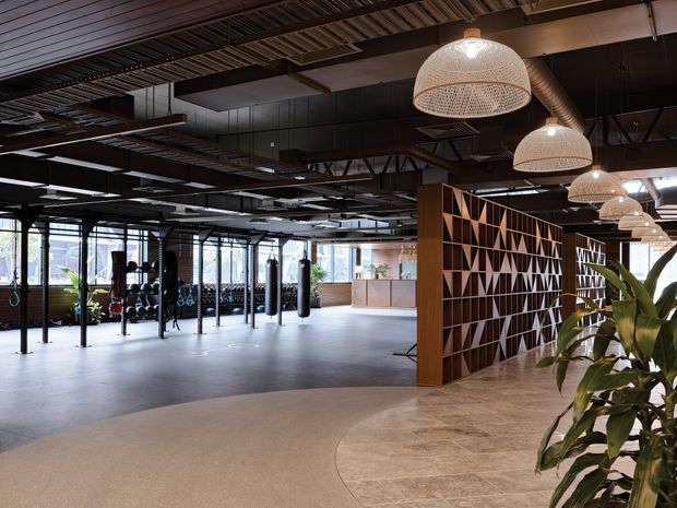 A spacious contemporary fitness studio with an open industrial-style ceiling, exposed beams and services, dark structural framing, large glazed windows, and a polished concrete-like floor. The space includes punching bags, exercise equipment, a reception counter, and a striking geometric timber privacy screen. Woven pendant lights and indoor plants add warmth to the otherwise raw, modern commercial interior.