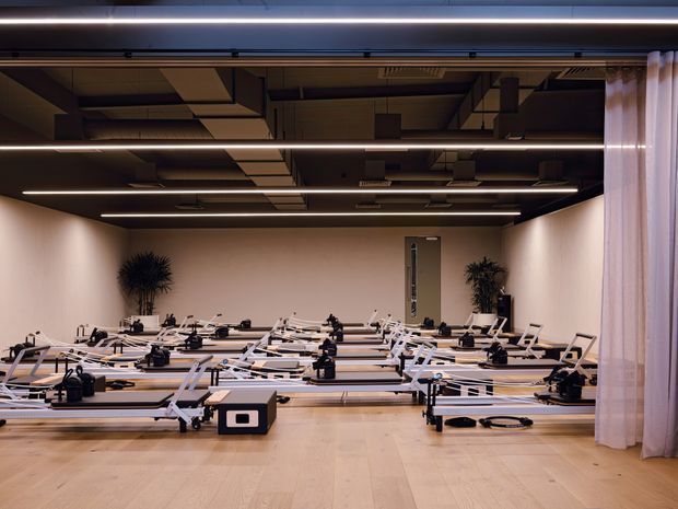 A spacious Pilates studio with multiple reformer machines arranged in rows on a pale timber floor. The room has warm beige walls, a dark exposed services ceiling with long linear strip lighting, full-height curtains along one side, and sparse potted greenery.