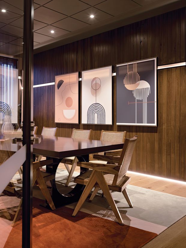 A sophisticated dining room with a large dark timber dining table surrounded by woven cane and timber chairs. The room features rich vertical dark wood wall panelling, warm integrated floor-level lighting, framed geometric artwork, a tiled acoustic ceiling, and layered neutral and rust-toned rugs. A glass partition or reflective surface is visible in the foreground.