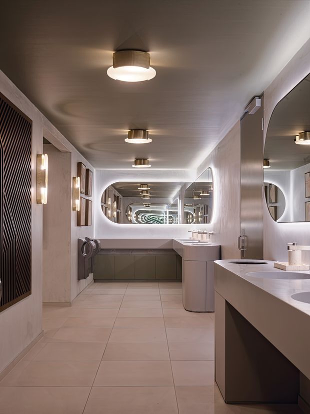 A long, refined contemporary bathroom with multiple vanity basins, extensive mirrors creating a repeating corridor effect, pale large-format tiled flooring, warm taupe wall finishes, rounded architectural openings, integrated cabinetry, and layered cylindrical ceiling and wall lighting. The space has a softly luxurious, symmetrical, hotel-like atmosphere.