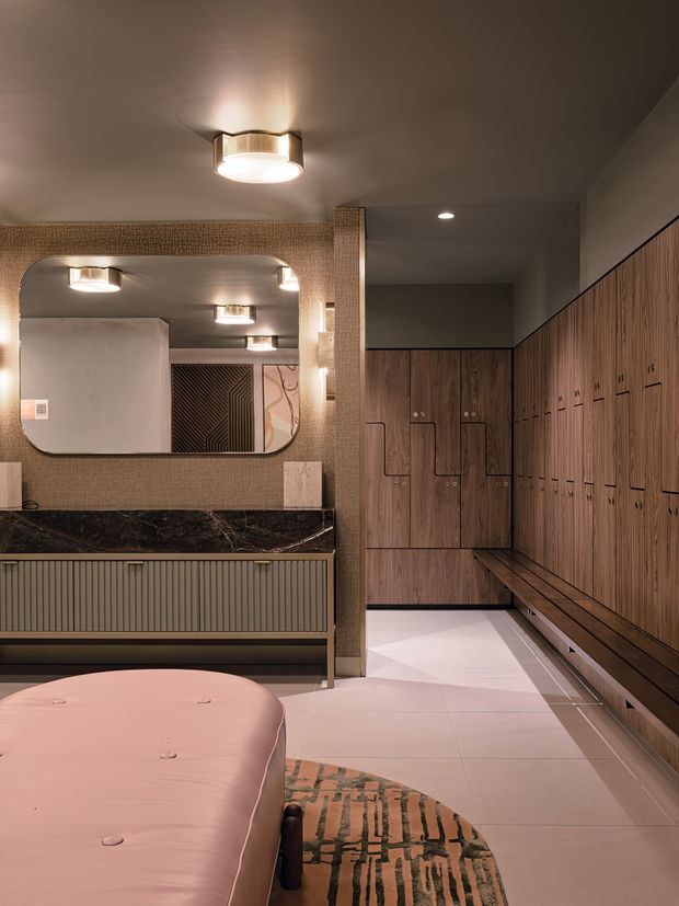 A refined modern locker-room or changing area with extensive warm wood locker cabinetry, an upholstered pink curved bench in the foreground, a dark stone vanity counter, ribbed cabinetry, textured wall tiles, rounded mirror, and warm integrated and ceiling lighting. The space has a sophisticated boutique wellness or hospitality feel.
