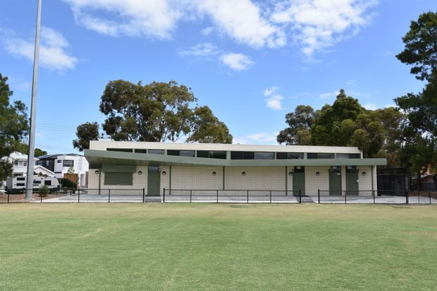 A low-rise institutional or commercial building viewed from across a broad grassed field. The long, rectilinear facade has pale masonry walls, a shallow low-pitched roof with a continuous clerestory-style strip of glazing, repeated green doors and shutters, and a black metal fence. Mature trees and neighbouring buildings are visible in the background under a bright blue sky.