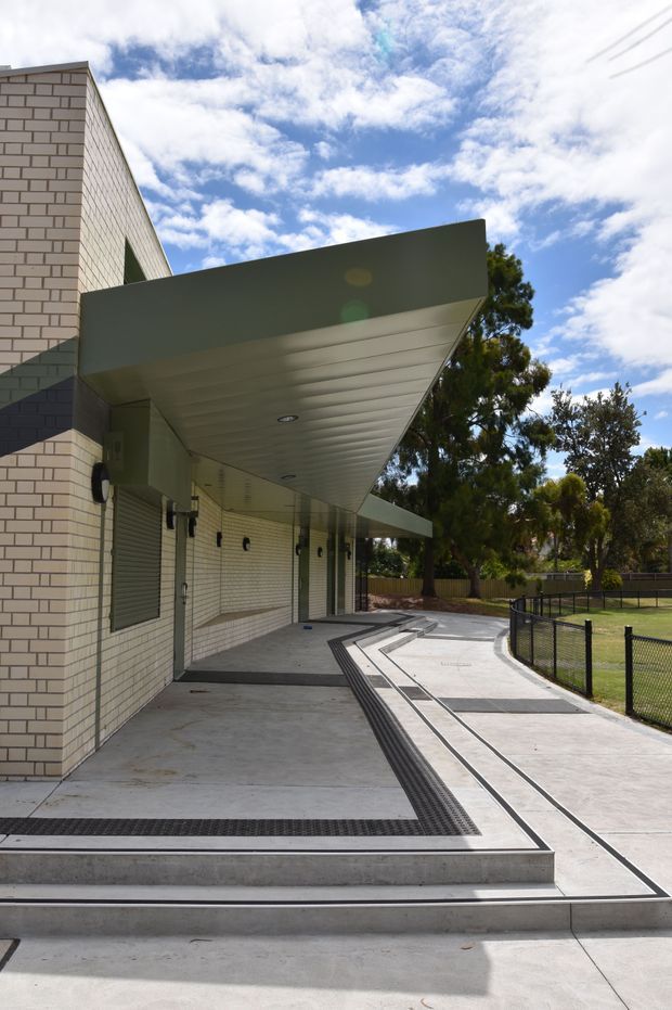 Exterior view of a contemporary institutional or commercial building entrance, featuring pale brick walls, a broad projecting metal canopy with recessed lights, roller shutters, accessible paved pathways with tactile drainage strips, and a fenced landscaped area beside the building. The image is photographed outdoors under a bright partly cloudy sky.