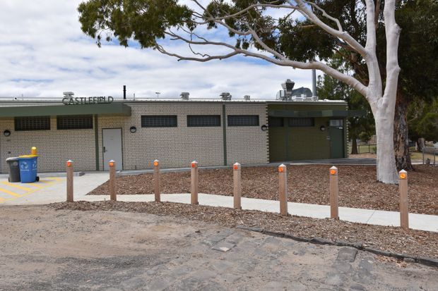 Single-storey institutional or commercial brick building viewed from outside, with a low flat roof, barred horizontal windows, plain service doors, rooftop mechanical equipment, bollards, paved access paths, and a large eucalyptus tree in the foreground. The facade is utilitarian and understated, with muted grey, beige and green tones.
