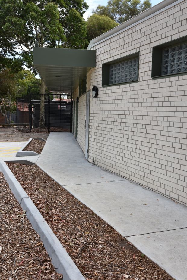 Exterior view of a low-rise building with a pale brick facade, dark-framed glass-block windows, a projecting flat-roof canopy, and a concrete pedestrian path beside landscaped mulch beds. A black metal security gate and covered entry are visible at the rear.