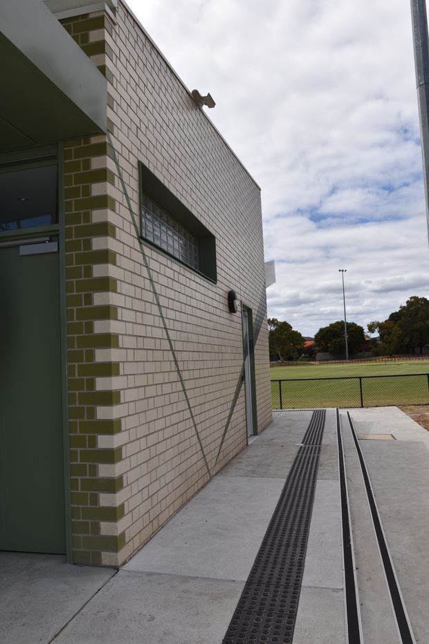Exterior view of a contemporary institutional or sports facility beside a fenced grass field. The building has pale cream brickwork with olive-green brick accents, a recessed glass-block window, dark green metal doors, and a concrete accessible walkway with linear drainage grates and tactile guidance strips. Overcast sky and mature trees are visible in the background.