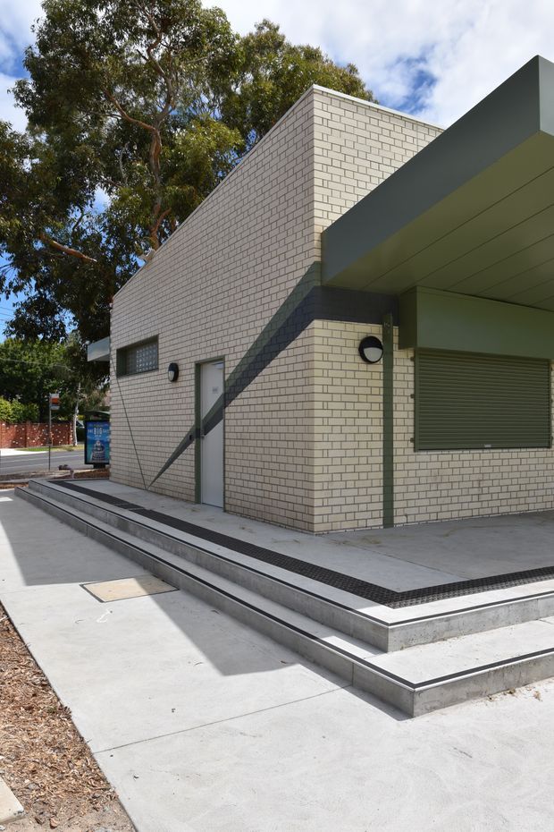 Exterior view of a compact contemporary building with pale cream brick walls, a projecting dark grey canopy, roller-shuttered opening, narrow windows, and raised concrete paving with linear drainage grilles. Mature trees and a suburban street are visible around the structure.