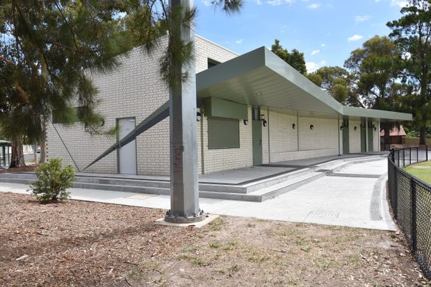 Exterior view of a low-rise institutional or commercial building with pale brick walls, a broad projecting metal canopy, recessed entrances, small circular wall lights, concrete steps and accessible paving. A fenced landscaped verge and mature trees surround the modern building.