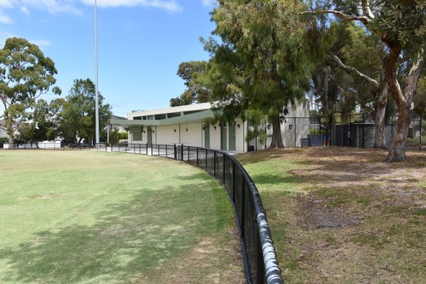 A low-rise modern building beside a broad maintained grassy field, viewed from outdoors. The building has a long pale rendered or masonry facade, narrow horizontal clerestory-style windows, simple projecting roof forms, dark-framed doors and windows, and a black metal security or boundary fence curving along the field. Mature eucalyptus-like trees surround the site under a clear blue sky.