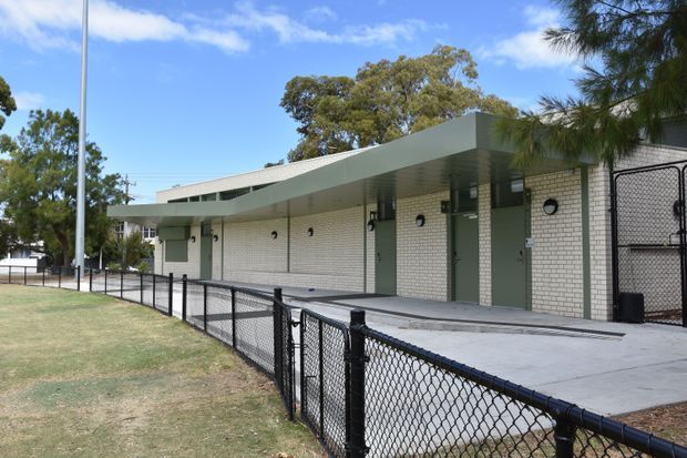Exterior view of a low-rise institutional or commercial building with a long, shallow flat roof canopy, pale painted brick walls, regularly spaced dark-framed doors and small circular wall lights. A paved walkway runs alongside a black chain-link fence bordering a landscaped grass area. The image is photographed outdoors under a bright blue sky.
