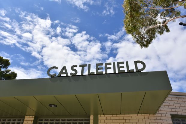 Exterior architectural photograph of a low-rise commercial or institutional building entrance, featuring a broad flat projecting canopy with large three-dimensional CASTLEFIELD lettering, pale brick walling, recessed downlights, and glazed blockwork beneath. The image is viewed from below against a blue sky with trees framing the building.