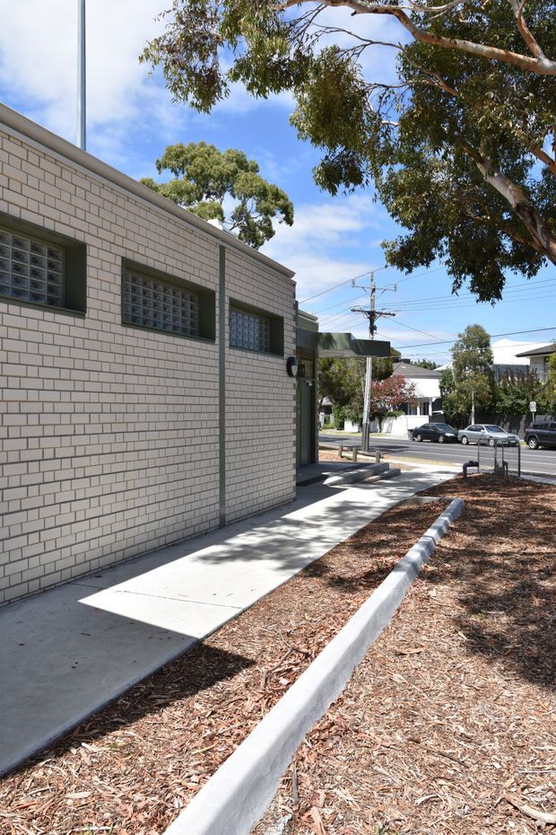 Exterior view of a low-rise light brick building beside a concrete footpath and landscaped verge. The facade features horizontal brickwork, dark-framed glass-block windows, and a simple flat or low-pitched roof edge. Mature eucalyptus trees, suburban streetscape, utility lines, and neighbouring buildings are visible under bright daylight.