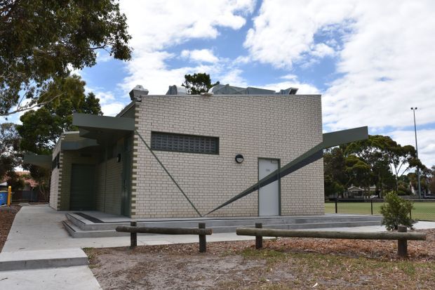 Exterior view of a low-rise contemporary brick building in a landscaped recreational setting. The structure has pale cream brickwork, dark grey angular projecting roof or canopy forms, a narrow horizontal glazed-block window, a simple metal entry door, and concrete paving around the perimeter. Timber post-and-rail fencing and mature trees are visible in the foreground and background.