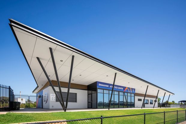 Modern single-storey athletics centre viewed from outside, featuring a dramatic projecting flat canopy with sharply angled dark steel support columns, white soffit panels, timber-look ceiling accent, glazed entrance frontage, and rendered light-coloured walls. The building sits beside fenced sports grounds under a clear blue sky.