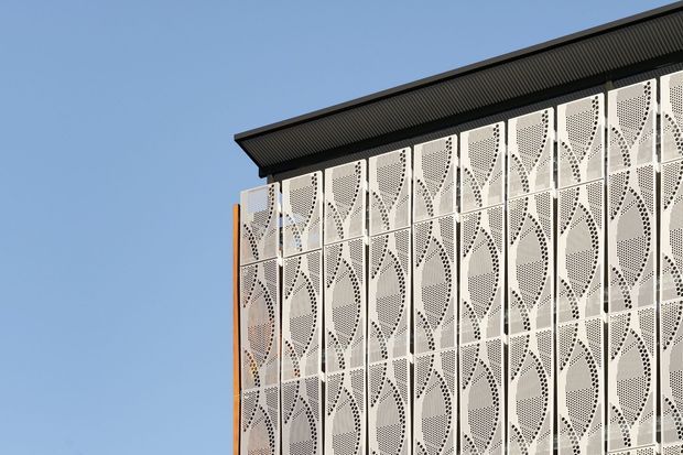 Close-up exterior view of a contemporary building facade beneath a dark, shallow sloping roof. The facade is clad in repeated white perforated metal panels with an intricate geometric pattern, creating a decorative screened elevation against a clear blue sky.