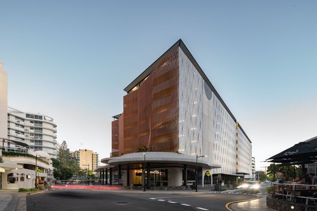 Urban architectural exterior of a contemporary multi-storey commercial building on a street corner. The angular building has a sharply pitched triangular roofline, a warm rust-coloured vertical screened facade on one side, and a pale translucent or patterned facade on the other. The ground level contains glazed retail or hospitality frontage beneath a broad curved canopy. Surrounding apartment and commercial buildings, street paving, signage, and outdoor cafe seating reinforce the dense urban setting.