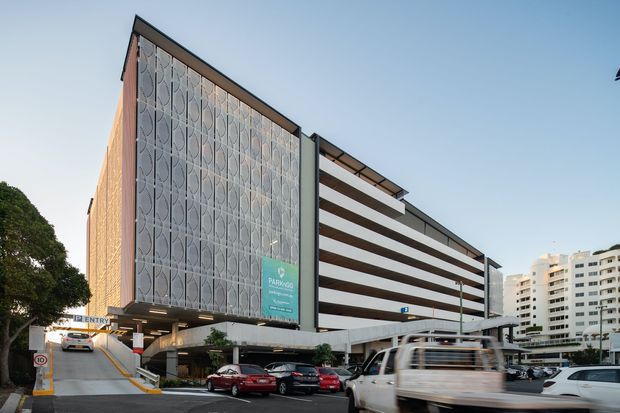 Exterior view of a multi-storey urban parking garage with open horizontal parking levels and a distinctive patterned translucent facade screen. The structure includes a vehicle entry ramp, ground-level parking, signage, and surrounding high-rise buildings.