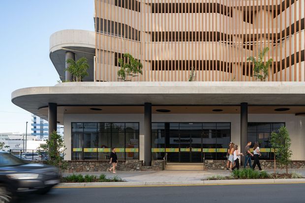 Exterior street-level view of a contemporary multi-storey building with rounded balconies and a prominent vertical timber-look screening facade. The ground floor has large glazed commercial or lobby spaces, a broad concrete canopy supported by dark columns, stone-clad plinths, landscaping, pedestrians, and a roadway in front.