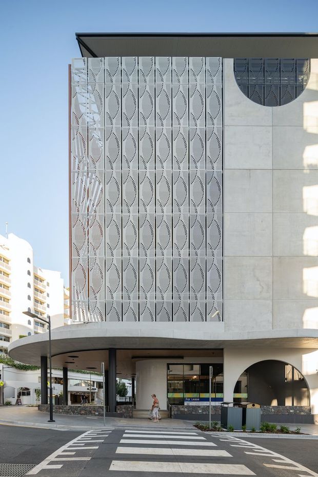 Street-level exterior view of a contemporary multi-storey commercial building in an urban setting. The facade combines a perforated decorative metal screen with a repeating organic pattern, large pale concrete or stone panels, curved arched openings, and a broad rounded canopy over the ground-floor entrance. A zebra crossing and neighbouring apartment buildings are visible in the foreground and background.