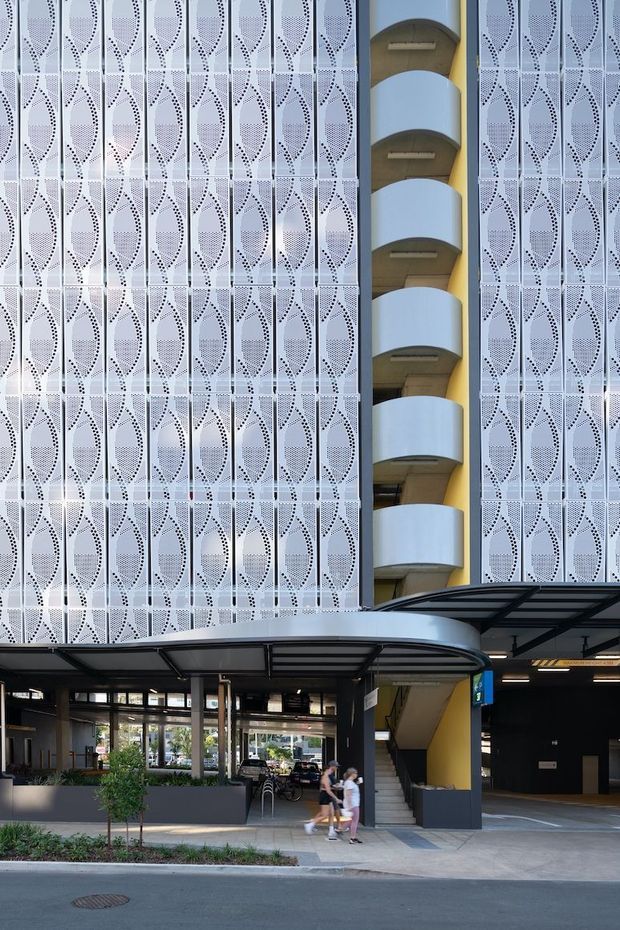 A contemporary multi-storey commercial building facade viewed from the street. The building features extensive white perforated metal screening with a repeating organic geometric pattern, a narrow vertical stairwell or balcony void with curved white projecting slabs and yellow internal walls, and a dark steel-and-glass entrance canopy. The ground level contains a cafe or lobby-like glazed space, planter beds, an external stair, and a covered vehicle entry or parking area.