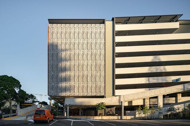 Exterior photograph of a contemporary multi-storey commercial building, likely a parking or mixed-use structure. The facade combines a tall decorative perforated or patterned screen with a horizontally banded open facade, supported by exposed concrete elements and ground-level columns. The image is taken from a street-level parking area under clear daylight.