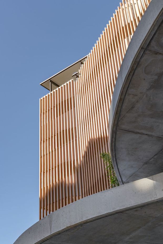 Upward-looking architectural exterior view showing a contemporary multi-level building with a tall vertical timber-slat screen facade, exposed concrete curved balcony or walkway structures, and a projecting roof canopy against a clear blue sky.