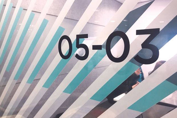 Close-up view of a contemporary graphic glass or glazed partition featuring large black “05–03” numerals over diagonal white, grey, turquoise, and dark geometric stripes. A person and interior space are partially visible behind the patterned surface.