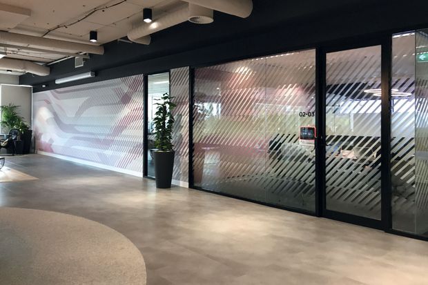 A modern commercial interior corridor or lobby with polished concrete flooring, black-framed glazed office partitions and doors featuring diagonal frosted privacy bands, exposed services and ductwork in the ceiling, and a large abstract graphic wall mural. Potted plants add greenery to the otherwise industrial office environment.