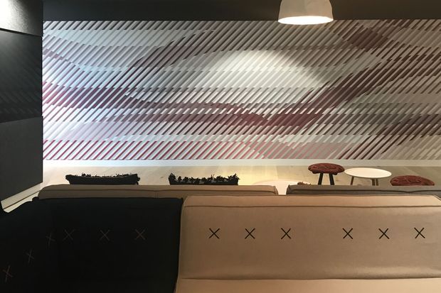 A dimly lit contemporary interior featuring a large upholstered sectional sofa in the foreground, with a dramatic textured diagonal-slat wall or glass partition behind it. The space includes a warm pendant light, low tables, and subtle decorative planting, creating a refined lounge or reception setting.