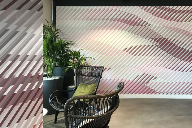 A contemporary interior seating area with two black woven lounge chairs, a small round table, and potted plants. The space is defined by a large geometric wall feature composed of diagonal burgundy, grey, white, and pink stripes, with a smooth grey floor and dark framing elements.