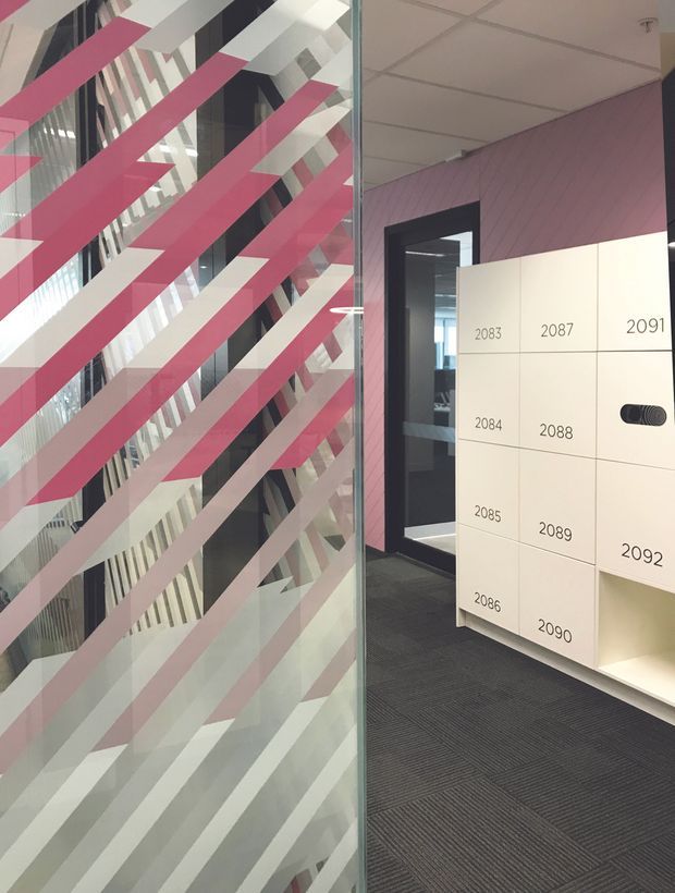 A contemporary commercial interior lobby or circulation area with a large glass partition decorated in bold diagonal pink, white and grey stripes. A wall-mounted white numbered locker or mail storage unit is positioned beside a dark-framed glazed doorway. The space has pink vertical wall panelling, a suspended acoustic tile ceiling, and dark carpet tiles.