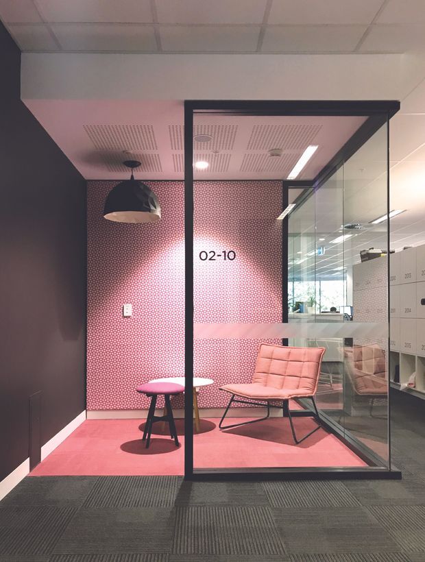 A compact enclosed office or meeting booth within a commercial interior, defined by black-framed glass walls. The space has a pink patterned feature wall, pink carpet, a sculptural dark pendant light, a lounge chair, and a small round table. Grey carpet tiles and office glazing are visible outside the booth, with a reflected corridor and workspaces beyond.