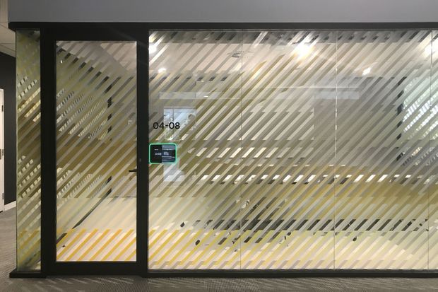 A glazed commercial office or meeting-room frontage viewed from a corridor, with dark metal framing and extensive glass panels decorated with diagonal frosted stripes. A glass door includes access-control hardware and room numbering, while desks and office furnishings are faintly visible through the patterned glazing. The scene has a contemporary, functional workplace character.